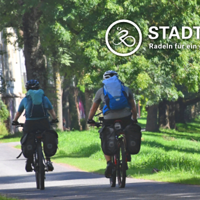 Zwei Fahrradfahrer mit Gepäck befahren einen Radweg mit Alleebäumen. Im Bild befindet sich eine Grafik mit dem weißen Schriftzug "Stadtradeln" und Logo eines stilisierten Radfahrers